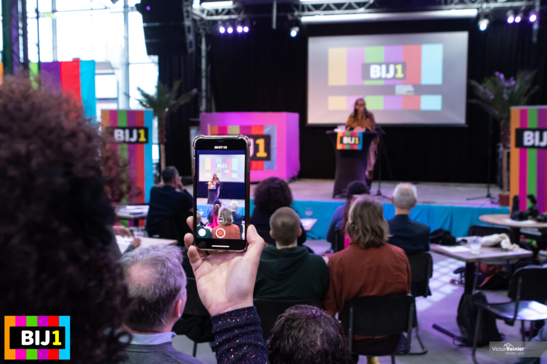 Een foto van het BIJ1 podium tijdens de kick off op 2 februari 2020. Veel BIJ1 logo's. Professor Gloria Wekker staat op het podium te spreken. Iemand filmt haar met een mobiele telefoon.