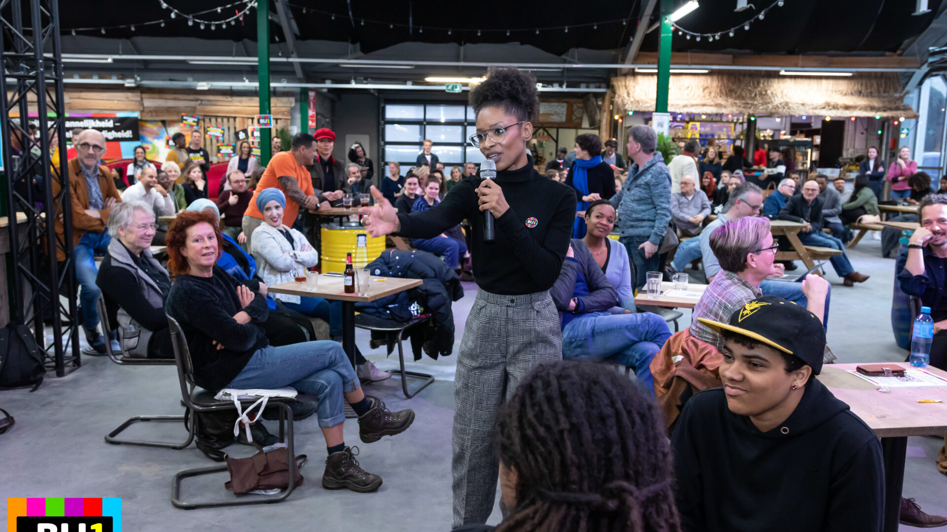 Sylvana Simons, zwarte vrouw met haar krullende haar opgestoken, spreekt in een microfoon die ze vasthoudt, terwjl ze haar andere hand uitnodigend uitsteekt. Ze staat temidden van een ruimte waar veel mensen zitten aan tafeltjes. Het ziet er feestelijk uit.
