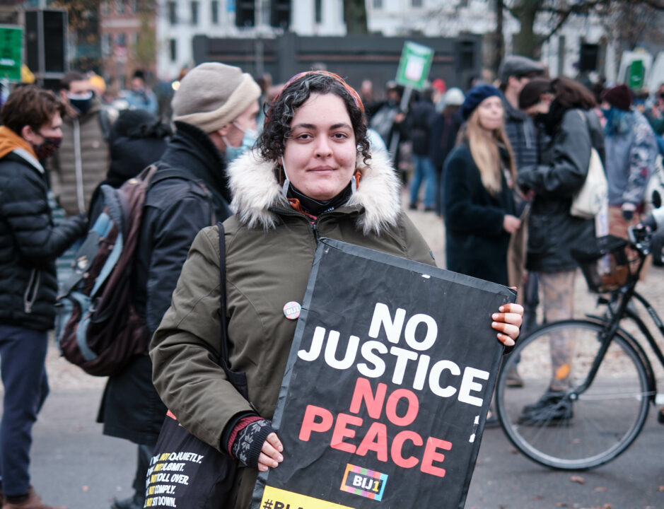 Een lichtbruine vrouw met krullen kijkt de camera in en houdt een bord vast waarop staat "No Justice No Peace" en het BIJ1 logo. Op de achtergrond zijn meer mensen te zien.