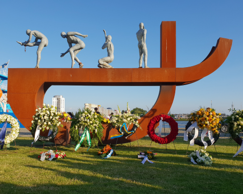 Het nationaal slavernijmonument, een metershoge roestbruine balk met gebogen vorm eronder, met daarop vier menselijke figuren in diverse houdingen. Er staan grote bloemenkransen voor het monument.