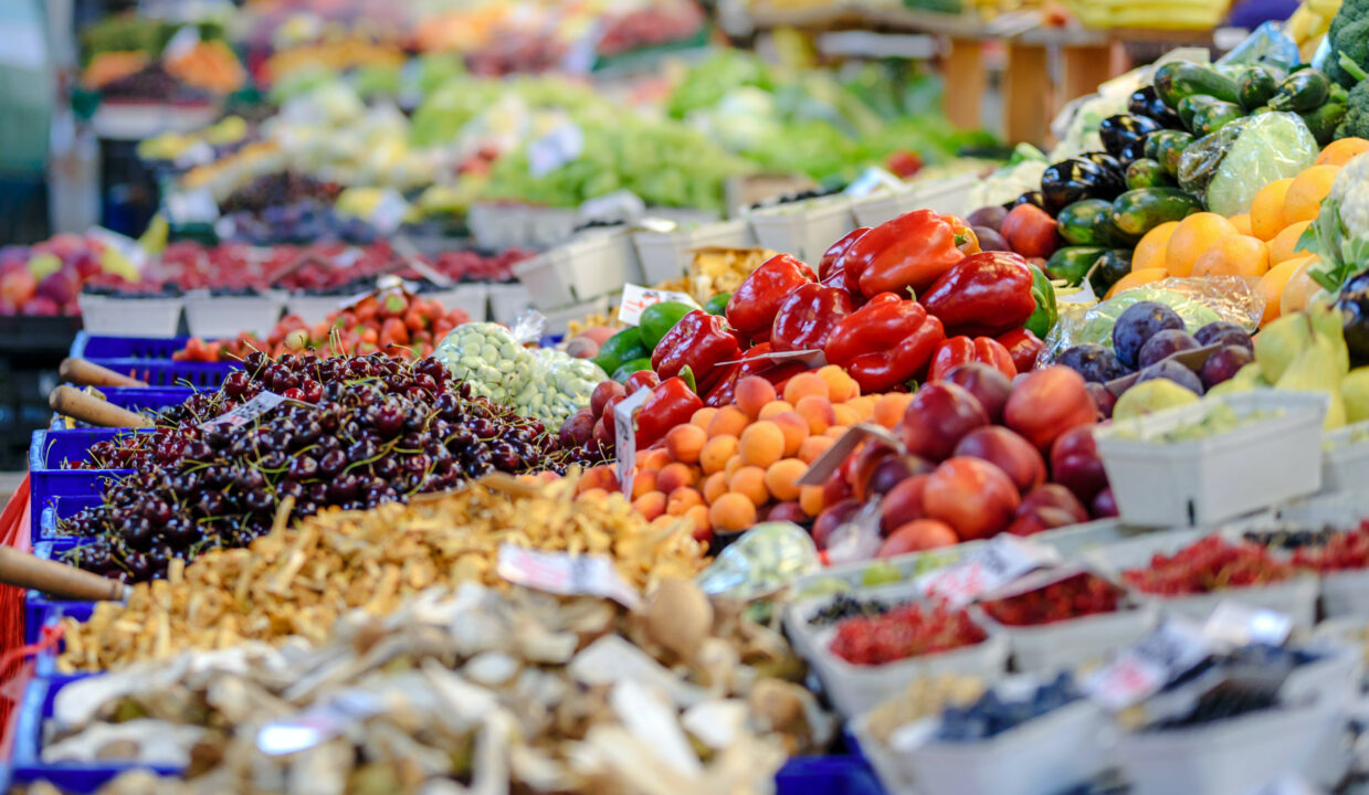 Op een uitgestalde marktkraam liggen allerlei soorten fruit en groenten: druiven, paprika, perzikken, aardappelen.