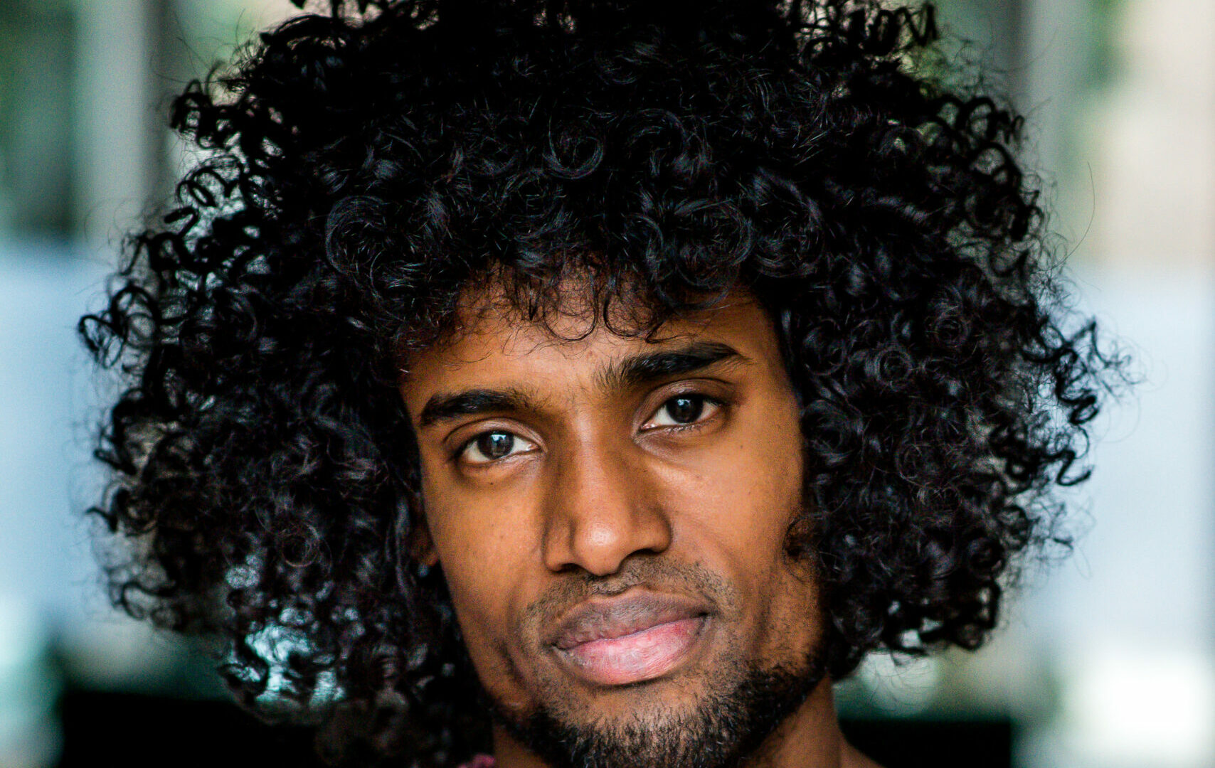 Portret van Daryll Ricardo Landbrug. Hij is een zwarte man met een sikje en een groot afro-kapsel. Hij draagt een roze trui en kijkt vriendelijk en serieus de camera in.
