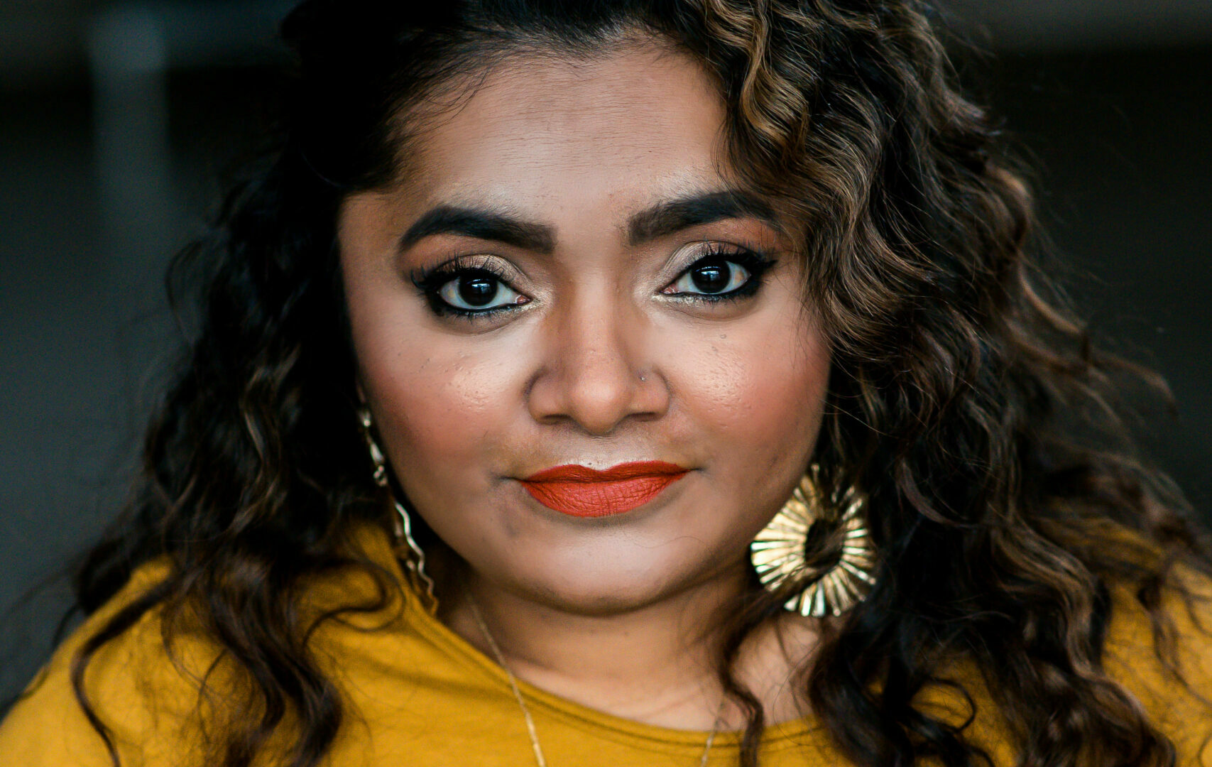 Portret van Jeanette Chedda. Ze kijkt recht in de camera. Ze is een bruine vrouw met donkerbruin haar met lichtere highlights en krullen erin. Ze draagt lippenstift, grote gouden oorbellen en een okergele trui. Om haar nek een kettinkje waar Jeanette op staat.