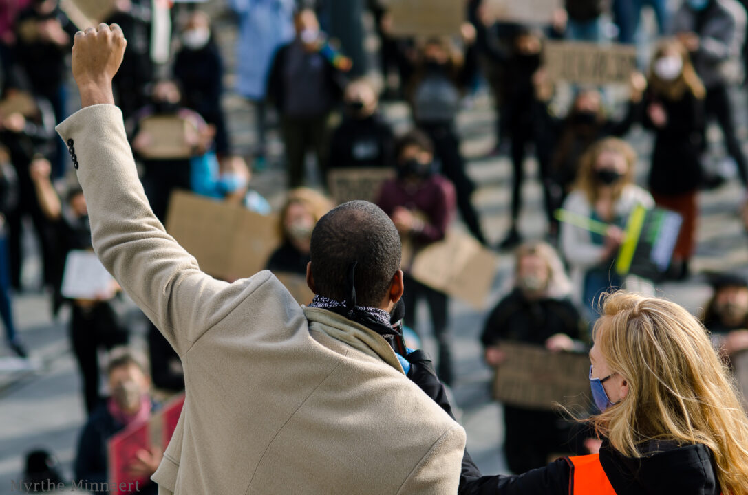 We zien de achterkant van een persoon die bij een protest zijn vuist omhoog steekt. Hij staat op een verhoging. Er staan protestanten naar hem te kijken die kartonnen bordjes met Black Lives Matter leuzen omhoog houden.