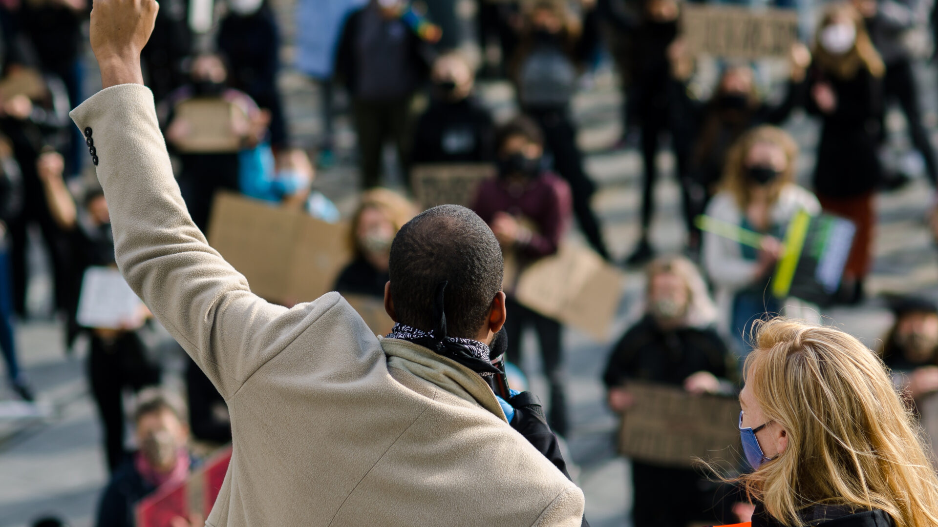 We zien de achterkant van een persoon die bij een protest zijn vuist omhoog steekt. Hij staat op een verhoging. Er staan protestanten naar hem te kijken die kartonnen bordjes met Black Lives Matter leuzen omhoog houden.