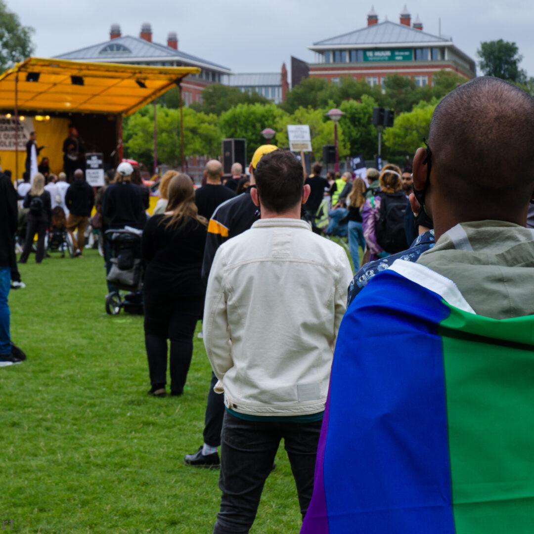 Een zwarte man heeft een regenboogvlag om zijn schouders. Hij staat tijdens ene protest op een grasveld naar het podium te kijken. Voor hem staan meer demonstranten op anderhalve meter afstand van elkaar