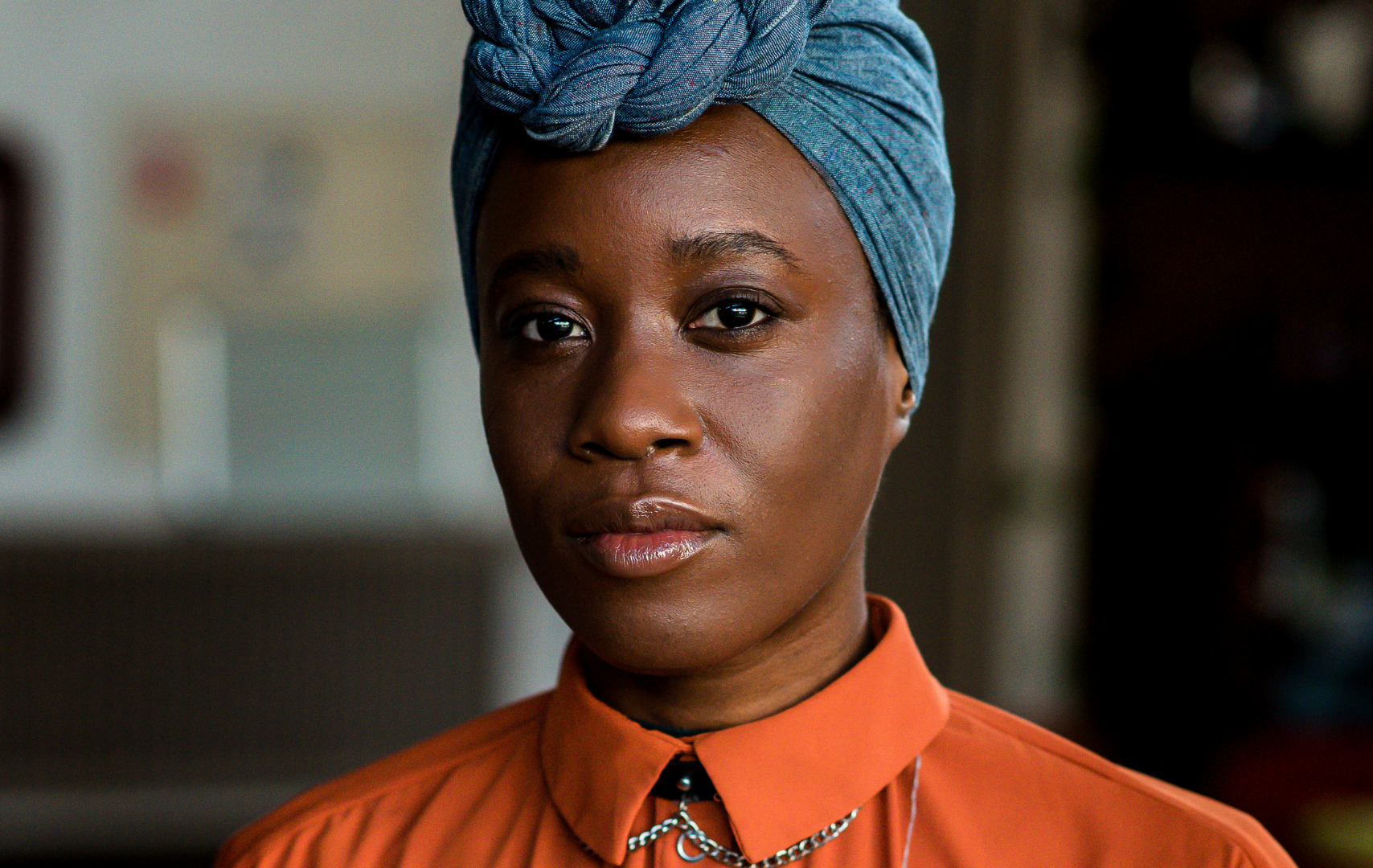 Portretfoto van Michantely de Jong. Een zwarte vrouw met een blauwe hoofddoek op. Ze draagt een oranje blouse.