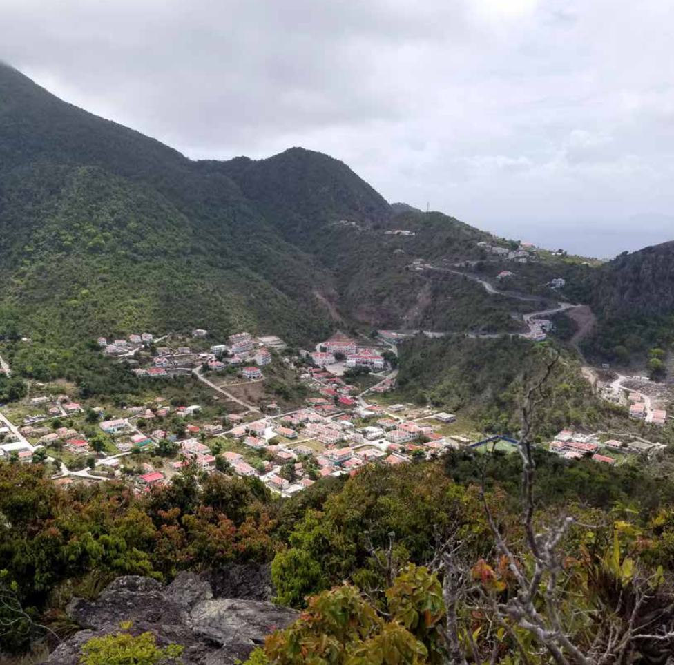 Een berglandschap, vanaf de hoogte gefotografeer. We zien tussen de heuvels een cluster aan huizen liggen. Dit is het eiland Saba