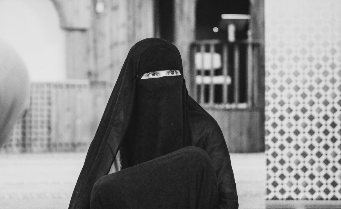 Zwart-wit foto van een vrouw in niqab. Op de achtergrond een hekje en een muur met tegeltjes erop.