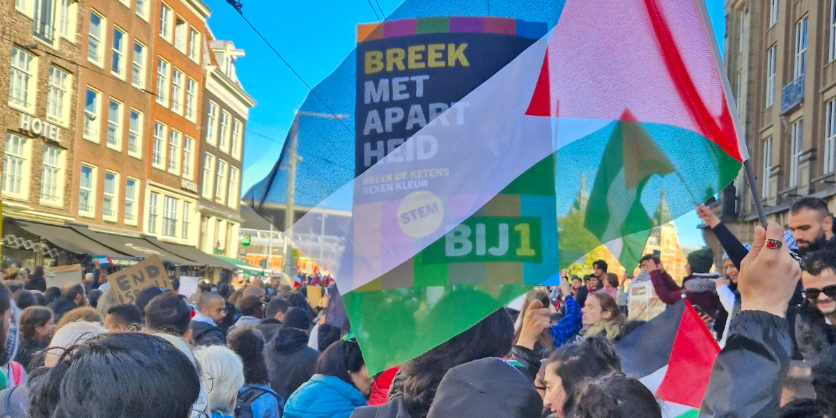 BIJ1-bord ‘Breek met apartheid’ is zichtbaar door transparante Palestijnse vlag tijdens demonstratie