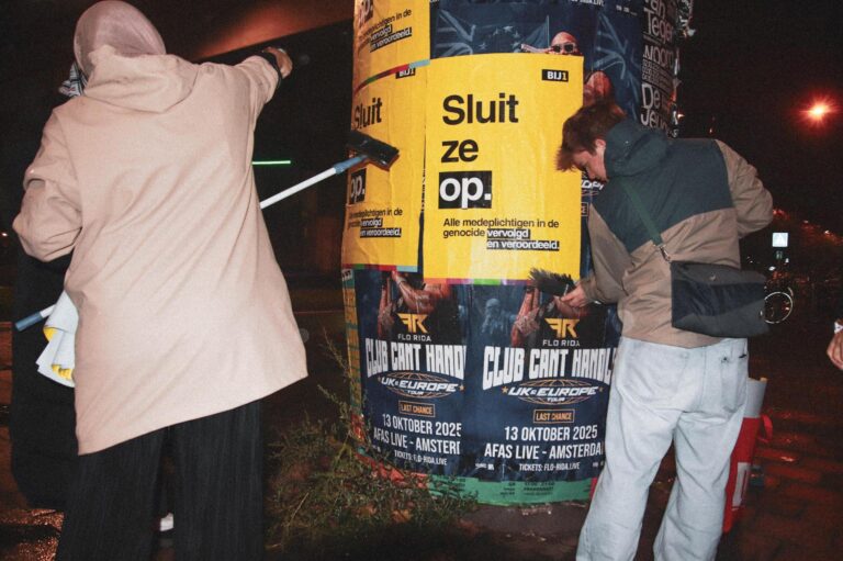 twee mensen plakken een poster van BIJ1 met de volgende tekst: Sluit. Ze. Op. Alle medeplichtigen in de genocide vervolgd en veroordeeld